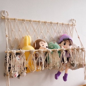 May include: A cream-coloured macrame wall hanging storage hammock featuring a wooden dowel frame and knotted cord netting. The hammock holds several plush toys, including a yellow rabbit, a monkey, and a doll wearing a purple hat and shirt.