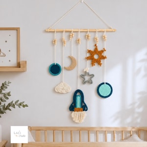 Könnte beinhalten: Ein Wandbehang mit einem Holzstab und daran befestigten weißen Schnüren. Die Schnüre halten verschiedene Formen, darunter einen dunkelblauen Kreis, eine beige Mondsichel, eine weiße Wolke, einen grauen Stern, eine senfgelbe Sonne und eine Rakete mit Fransen.