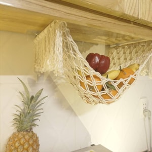 Op de afbeelding: Een witte katoenen touwhangmat die aan een houten kast hangt, gevuld met vers fruit, waaronder sinaasappelen, bananen en een rode paprika.