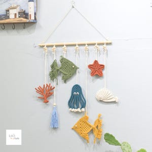 May include: Macrame wall hanging with sea creatures: a green fish, orange starfish, blue octopus, yellow fish, coral, and a seashell. The nautical decor is suspended from a wooden dowel with a triangle top and a blue tassel.