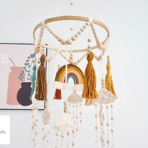 May include: A wooden baby mobile with a rainbow, clouds, and tassels hanging from a circular wooden frame. The mobile is decorated with wooden beads.