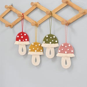 May include: Four handmade macrame mushroom ornaments in red, yellow, green, and pink, each with white polka dots and a cream-colored stem. They hang from a wooden rack against a light blue wall. The ornaments are a whimsical, decorative item.