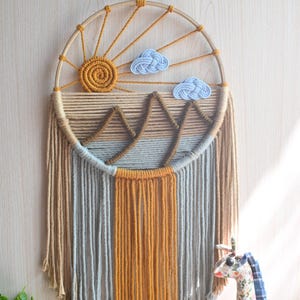 May include: Macrame wall hanging with a sun, clouds, and mountain design. The piece features a circular wooden frame with a woven sun and two blue clouds. The lower portion has vertical strands in shades of brown, gray, and orange. The text "Style B" is at the top.