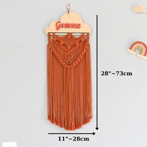 May include: A macrame wall hanging with a wooden cloud cutout with the name "Gemma" in red letters. The hanging is 28 inches tall and 11 inches wide.