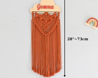 May include: A macrame wall hanging with a wooden cloud cutout with the name "Gemma" in red letters. The hanging is 73 cm tall and 28 cm wide.