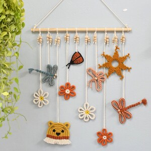 May include: A decorative wall hanging with crochet elements, such as flowers, dragonflies, a butterfly, and a bear. The piece is suspended from a wooden dowel with wooden beads and white cords. The color scheme includes orange, yellow, white, and blue, creating a cheerful aesthetic.