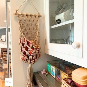 May include: A beige macrame hanging fruit basket made of knotted cord, suspended from a wooden dowel with a rope hanger. The basket features a diamond-patterned mesh design and a long tassel at the bottom, currently holding several red apples.