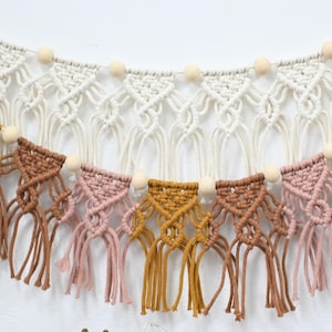 May include: A macrame wall hanging featuring a series of triangular knots in cream, dusty pink, mustard yellow, and terracotta brown cotton cord, accented with natural wooden beads and finished with long, loose fringe. Text reads Lee's Studio.