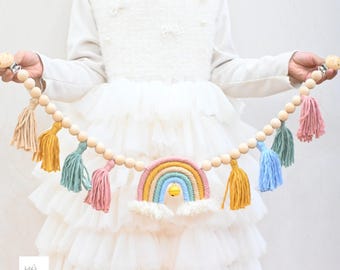 Pode incluir: Uma guirlanda decorativa com contas de madeira natural enfiadas junto a borlas de fio multicolorido em tons de rosa, amarelo, azul e verde. Um ornamento central de arco-íris em macramé com um pequeno sino amarelo pende do centro.
