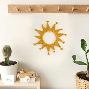 May include: A yellow macrame sun wall hanging with a circular centre and fringe details. The sun is hanging on a white wall with a wooden shelf below it.