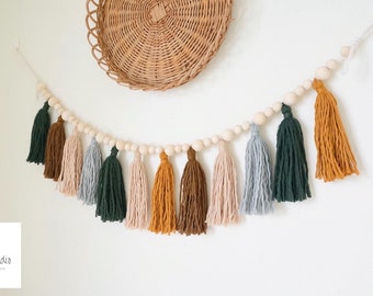 Può includere: Una ghirlanda con perline di legno e nappe di filato colorato in tonalità di verde, marrone, beige e grigio. La ghirlanda è appesa a un muro bianco.