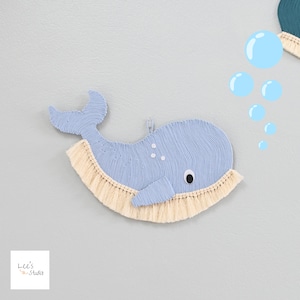 May include: A light blue whale-shaped wall hanging with a textured surface and a cream-colored fringe. The whale has a small eye and three white dots. Light blue bubbles are in the background. The logo "Lee's Studio" is in the bottom left corner.