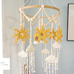 May include: A hanging mobile featuring a circular wooden frame adorned with natural wooden beads. Suspended from the frame are yellow macrame suns, white macrame clouds, and a small rainbow, all accented with white cord and wooden bead strands.