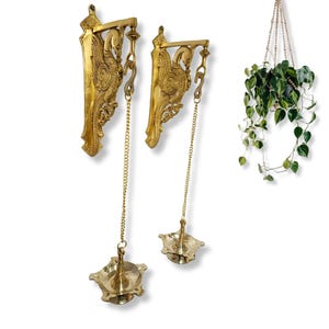 Peut inclure: Deux appliques murales dorées ornées d'un motif d'oiseau, suspendues par des chaînes. Une plante suspendue avec des feuilles vertes est également visible, ajoutant une touche naturelle.