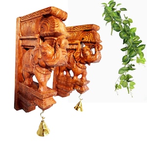 Może przedstawiać: Dwie misternie rzeźbione drewniane dekoracje ścienne ze słoniami. Słonie są brązowe, z szczegółowymi rzeźbieniami i złotymi dzwoneczkami wiszącymi poniżej. Po prawej stronie obrazu znajduje się zielony, liściasty pnącze.