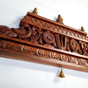 May include: A rectangular brown wooden wall hanging featuring intricate carvings of birds, floral patterns, and a central U-shaped symbol. Three small brass bells hang from the bottom edge, and three small brass finials are attached to the top edge.