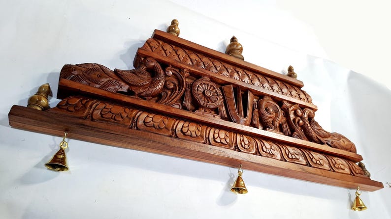 May include: A rectangular brown wooden wall hanging featuring intricate carvings of birds, floral patterns, and a central U-shaped symbol. Three small brass bells hang from the bottom edge, and three small brass finials are attached to the top edge.
