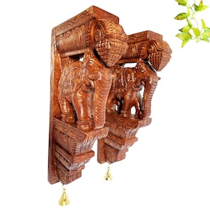 Puede incluir: Un par de soportes de pared de madera marrón con figuras de elefantes talladas a mano con detalles intrincados en los cuerpos y trompas. Cada soporte tiene una sección superior decorativa y una base escalonada con una pequeña campana de latón.