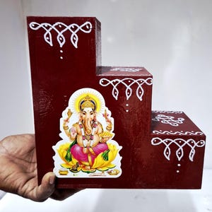 May include: A three-tiered, burgundy-colored wooden display stand. It features a colorful image of the Hindu deity Ganesha on the front. The stand is decorated with white painted designs. The stand is held by a person's hand.