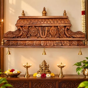 Hand carved wooden temple wall shelf with detailed peacock and floral carvings, three brass bells, and antique brown wood finish displayed in an elegant Indian pooja room with brass lamps and flowers.