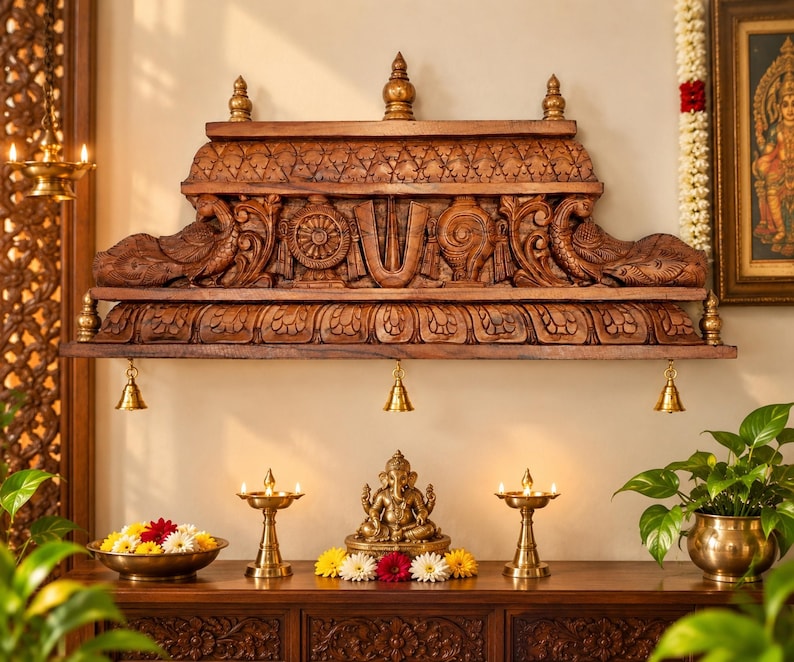 Hand carved wooden temple wall shelf with detailed peacock and floral carvings, three brass bells, and antique brown wood finish displayed in an elegant Indian pooja room with brass lamps and flowers.