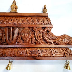May include: A rectangular brown wooden wall panel featuring intricate hand-carved details including a central U-shaped symbol, a wheel, a conch shell, and a peacock. The top and bottom edges are adorned with carved patterns and small brass bells.