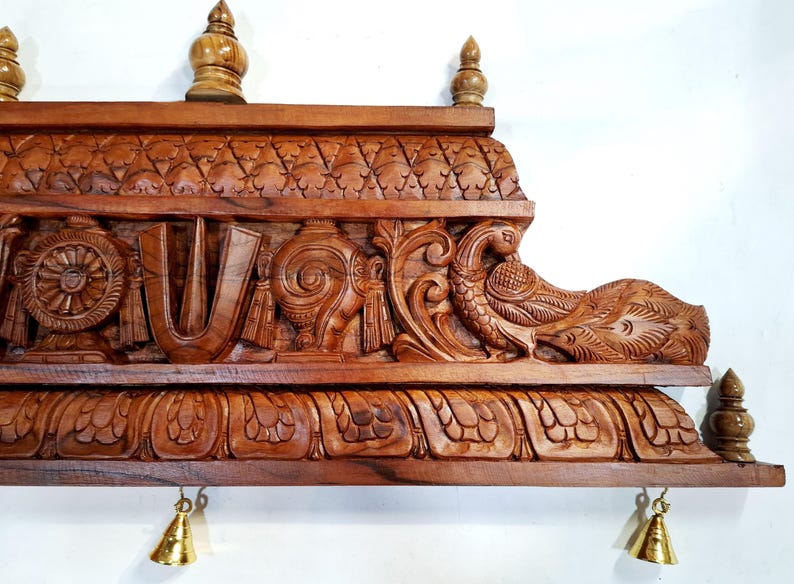 May include: A rectangular brown wooden wall panel featuring intricate hand-carved details including a central U-shaped symbol, a wheel, a conch shell, and a peacock. The top and bottom edges are adorned with carved patterns and small brass bells.
