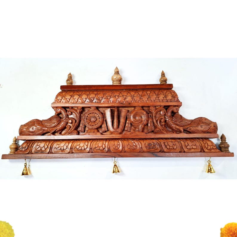 May include: A rectangular brown wooden wall hanging featuring intricate carvings of two peacocks, a central U-shaped symbol, a wheel, and a conch shell. The piece is topped with three small finials and has three small brass bells hanging from the bottom.