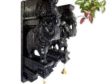 May include: Black wooden wall decor featuring carved elephants. The elephants stand on a decorative base, with gold-colored bells hanging below. The wall decor has an ornate, detailed design, and is suitable for home decor.