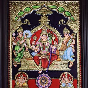 May include: A traditional Indian painting depicting a goddess seated on a throne with two female figures standing on either side. The painting is framed in a dark wood frame and features intricate gold details.