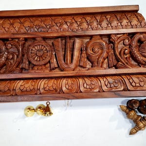May include: A rectangular brown wooden wall shelf featuring intricate hand-carved details including two peacocks, a central wheel, a conch shell, and a U-shaped symbol. The shelf includes small metal hooks and separate brass and wooden components.