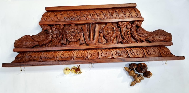 May include: A rectangular brown wooden wall shelf featuring intricate hand-carved details including two peacocks, a central wheel, a conch shell, and a U-shaped symbol. The shelf includes small metal hooks and separate brass and wooden components.