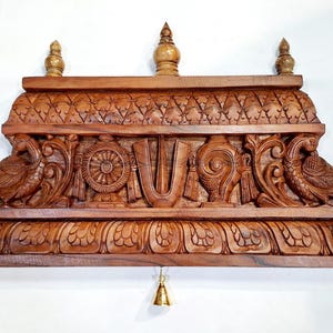 May include: A brown wooden wall shelf featuring intricate hand-carved details including two peacocks, a central U-shaped symbol, a wheel, and a conch shell. The shelf has a decorative top border, three small finials, and three hanging brass bells.