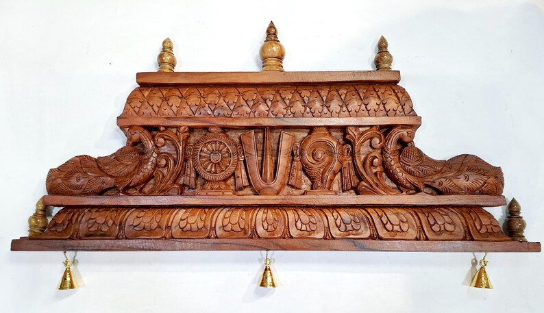 May include: A brown wooden wall shelf featuring intricate hand-carved details including two peacocks, a central U-shaped symbol, a wheel, and a conch shell. The shelf has a decorative top border, three small finials, and three hanging brass bells.
