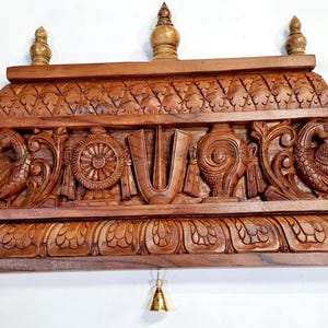 May include: A rectangular brown wooden wall shelf featuring intricate carvings of two peacocks, a central wheel, a conch shell, and a U-shaped symbol. The top and bottom edges have decorative floral patterns, and three small brass bells hang from the base.