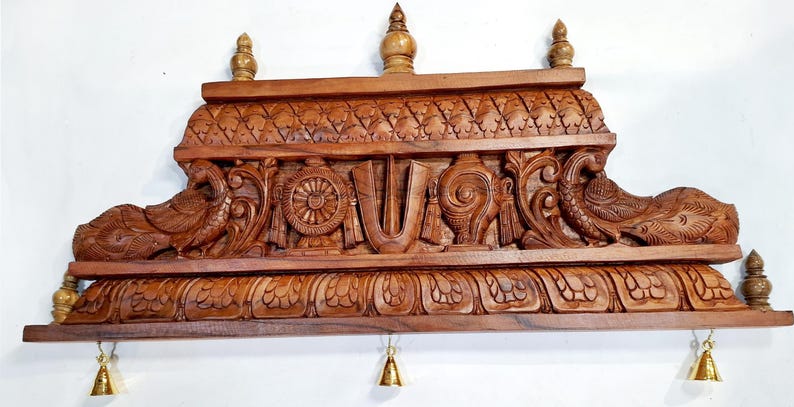 May include: A rectangular brown wooden wall shelf featuring intricate carvings of two peacocks, a central wheel, a conch shell, and a U-shaped symbol. The top and bottom edges have decorative floral patterns, and three small brass bells hang from the base.