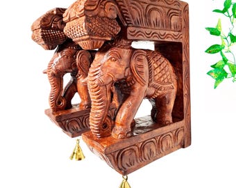 May include: A pair of brown wooden wall brackets featuring carved elephant figures with detailed textures and patterns. Each bracket has a small, polished gold-colored metal bell hanging from the bottom edge, suspended by a thin metal loop.