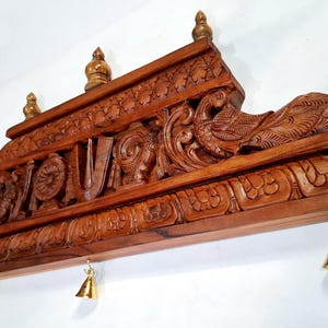 May include: A rectangular brown wooden wall hanging featuring intricate carvings of a peacock and traditional symbols. The top edge is adorned with three small wooden finials, while the bottom edge features three small hanging brass bells.