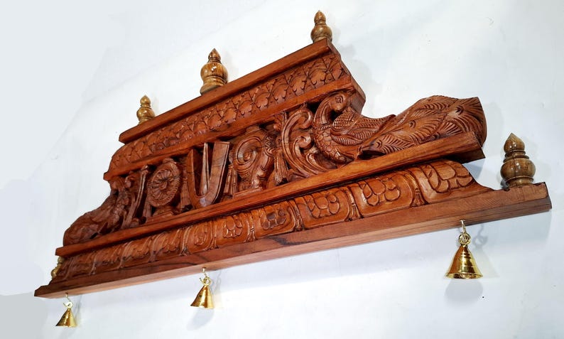 May include: A rectangular brown wooden wall hanging featuring intricate carvings of a peacock and traditional symbols. The top edge is adorned with three small wooden finials, while the bottom edge features three small hanging brass bells.