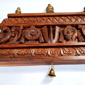 May include: A rectangular brown wooden wall hanging featuring intricate hand-carved details including a central U-shaped symbol, a wheel, a conch shell, and two birds on the sides. Three small wooden finials sit on top, and a brass bell hangs below.