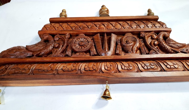 May include: A rectangular brown wooden wall hanging featuring intricate hand-carved details including a central U-shaped symbol, a wheel, a conch shell, and two birds on the sides. Three small wooden finials sit on top, and a brass bell hangs below.