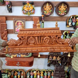 May include: A rectangular wooden temple structure featuring intricate hand-carved details, including two peacock figures on the sides, a central religious symbol, and a decorative roof. Small brass bells hang from the bottom edge of the frame.