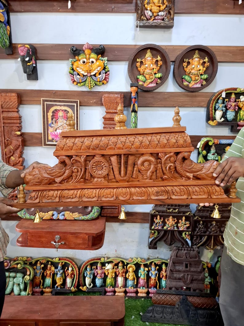 May include: A rectangular wooden temple structure featuring intricate hand-carved details, including two peacock figures on the sides, a central religious symbol, and a decorative roof. Small brass bells hang from the bottom edge of the frame.