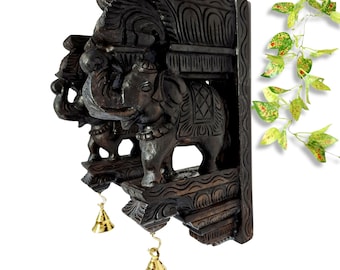 May include: Dark brown, intricately carved wooden wall hanging featuring three elephants. The elephants are adorned with detailed carvings and stand on a decorative platform. Two golden bells hang below. A decorative piece for home decor.