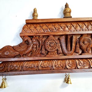 May include: A rectangular brown wooden wall shelf featuring intricate hand-carved details including a peacock, a wheel, a U-shaped symbol, and a conch shell. The shelf is topped with three small wooden finials and has two small brass bells hanging.