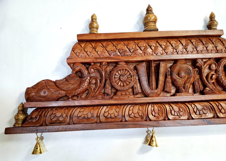 May include: A rectangular brown wooden wall shelf featuring intricate hand-carved details including a peacock, a wheel, a U-shaped symbol, and a conch shell. The shelf is topped with three small wooden finials and has two small brass bells hanging.