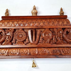 May include: A rectangular brown wooden wall shelf featuring intricate hand-carved details including two peacocks on the sides, a central U-shaped symbol, a wheel, and a conch shell. The shelf has a tiered top with three finials and three small brass bells.