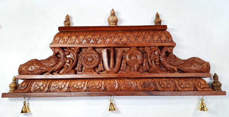 May include: A rectangular brown wooden wall shelf featuring intricate hand-carved details including two peacocks on the sides, a central U-shaped symbol, a wheel, and a conch shell. The shelf has a tiered top with three finials and three small brass bells.