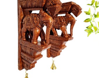 May include: A pair of brown wooden wall brackets featuring carved elephant figures and decorative patterns. Each bracket has a small gold-colored metal bell hanging from the bottom. The wood has a polished finish with detailed textures.