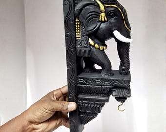 May include: Black wooden elephant wall shelf with gold and white accents. The elephant sculpture has detailed carvings and a hook for hanging items. The shelf is held by a hand.
